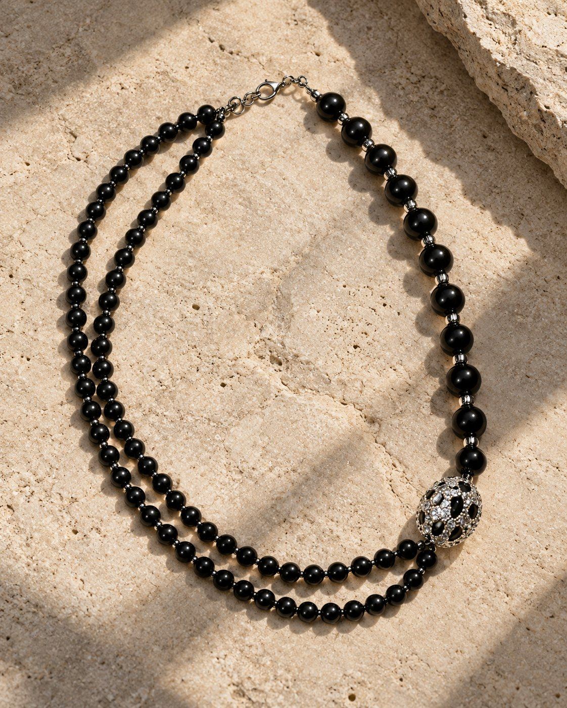 Mafdet necklace laid on travertine: two strands of glossy black beads joined by a silver-tone bead set with clear stones and black spots.