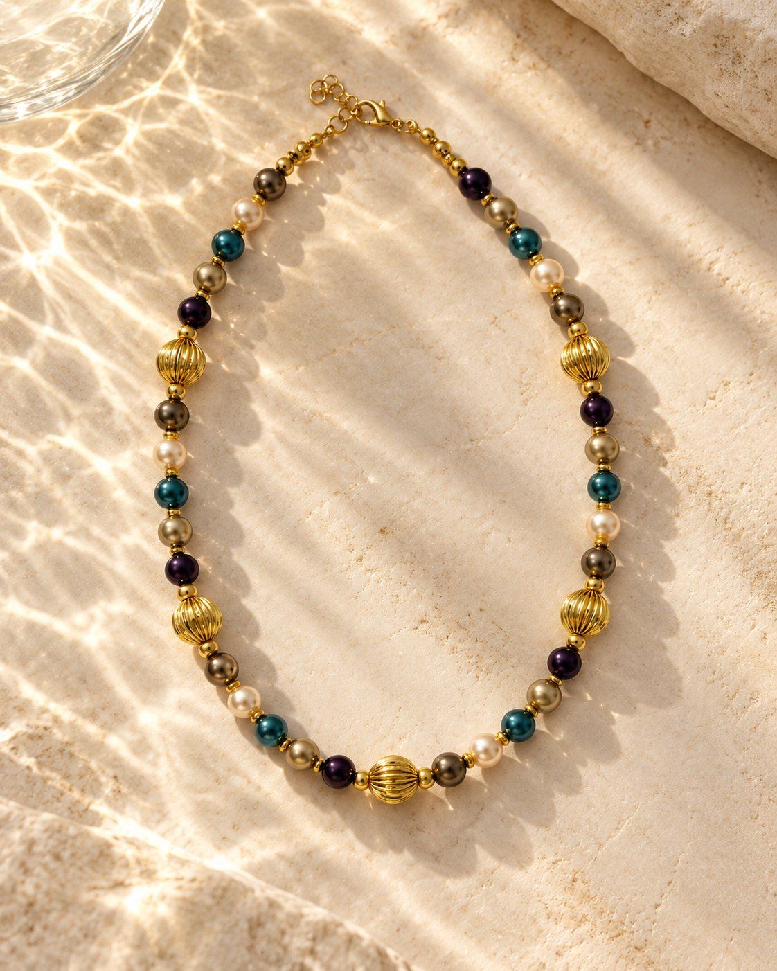Nanshe necklace laid on travertine in rippling water light: pearls in teal, aubergine, bronze and cream between fluted gold-tone beads.