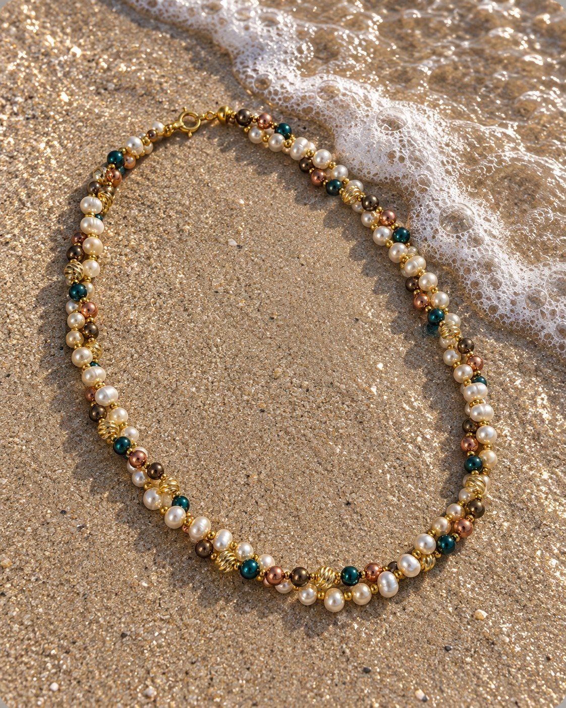 Phaenna necklace on wet sand at the water's edge: twisted strands of cream pearls with teal, bronze and rose-gold-tone beads.